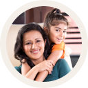 A woman wearing a green top holds a girl child on her back in a bright orange shirt, both smiling and enjoying.