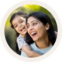 A woman holds a girl child on her back, both appearing joyful , with a greenery suggesting a sunny outdoor setting.