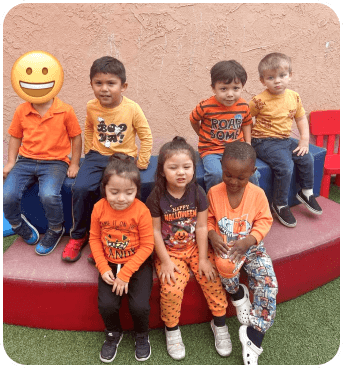 A group of seven children dressed in orange outfits, smiling and seated on a colorful play structure against a textured background.