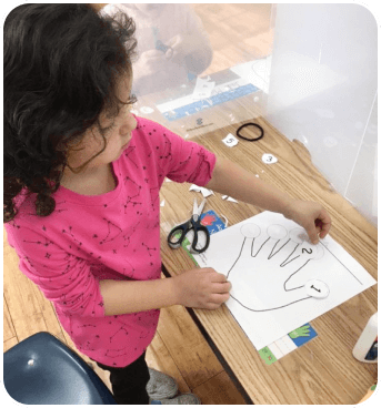 A child in a pink shirt decorates a hand-shaped paper with numbered circles while using scissors at a wooden table.