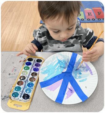 A child paints on a paper plate using watercolors, with blue tape creating sections for a colorful artwork.