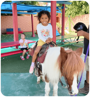 Two children are enjoying a pony ride at a playground, with colorful play equipment in the background and a caregiver nearby.