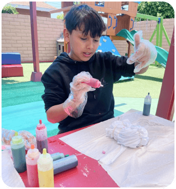 A child wearing gloves is using colorful squeeze bottles to dye a white cloth on a table in a bright outdoor setting with playground equipment.