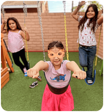 Three children are playing on swings in a vibrant playground, enjoying a sunny day with green grass underfoot.