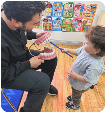 A person demonstrates dental care using a large model of teeth while a young child attentively holds a toothbrush, engaged in learning.