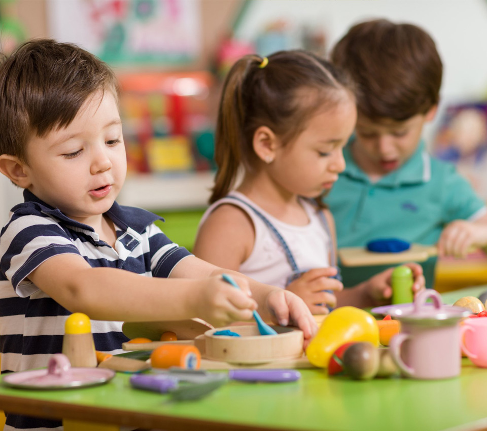 Children engage in creative play with colorful wooden toys and tools at a bright classroom table filled with playful materials.