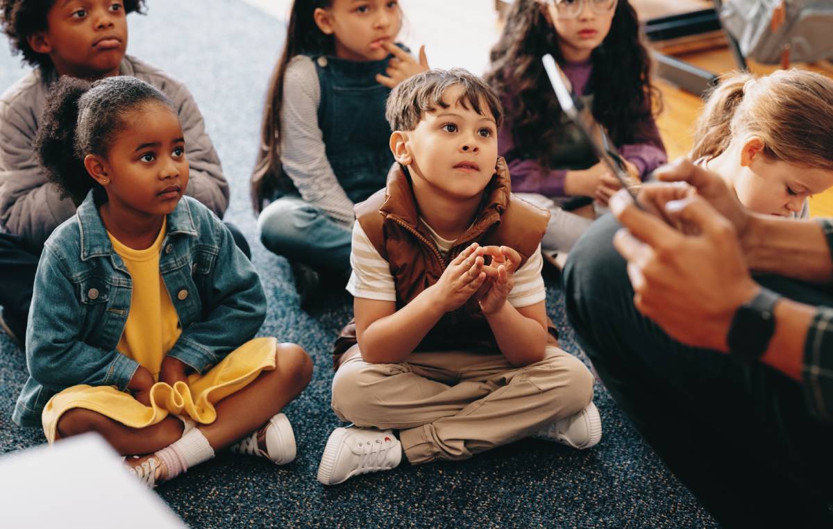Listening comprehension lesson in an elementary class. Kids pay attention to their teacher reading a story from a tablet. Group of diverse children sitting on the floor in a primary school.
